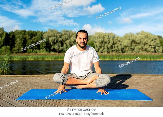 fitness, sport, yoga, people and healthy lifestyle concept - man making scale pose lotus variation on mat outdoors on river or lake berth