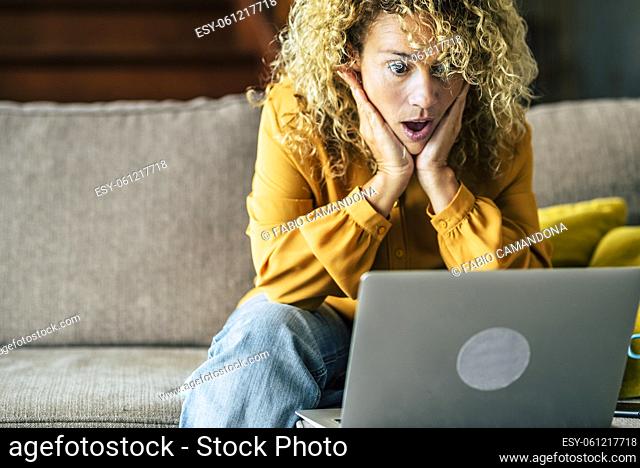 Happy smiling woman sitting at home on the sofa reading message looking at device screen using computer. Excited female feels satisfied positive emotions took...