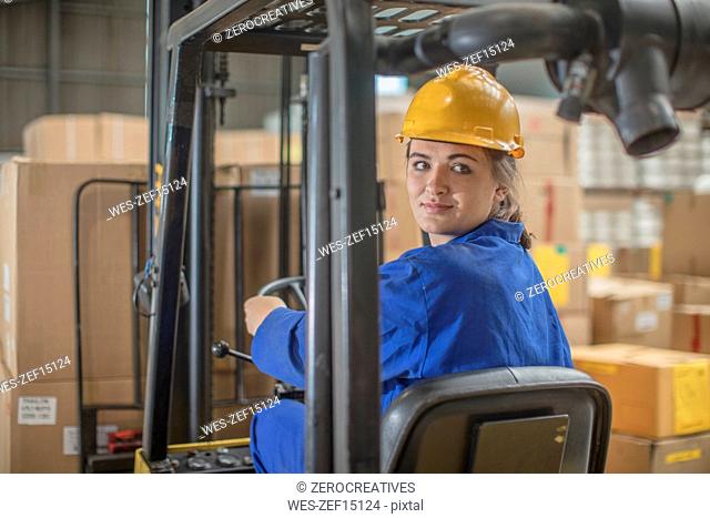 Smiling woman wearing hard hat driving forklift in factory