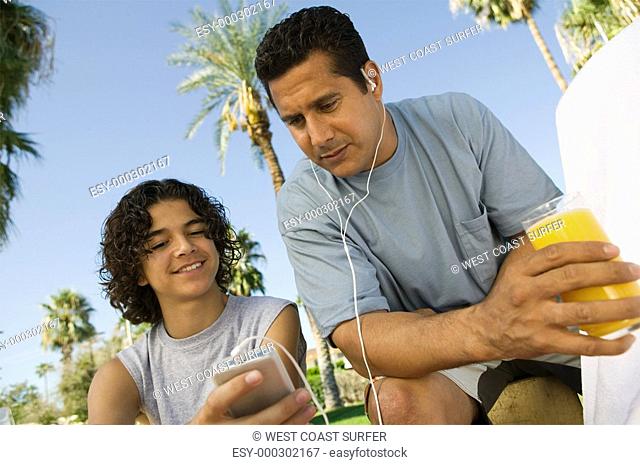 Boy 13-15 holding portable music player father listening with earphones and holding glass of juice