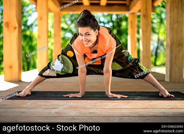 Handsome girl makes wide-legged forward bend on the black yoga mat on the wooden terrace on the nature background. She holds her hands on the mat