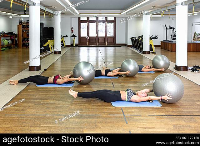 Group of barefoot sportswomen in activewear lying on floor and exercising with fit balls together during fitness training in spacious light gym