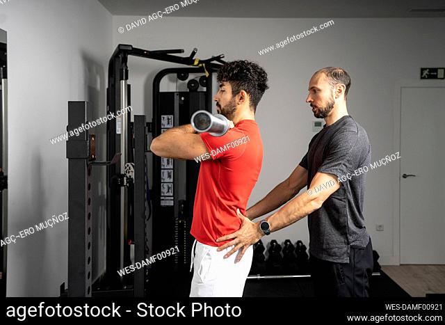 Instructor giving support to young man lifting barbell at gym