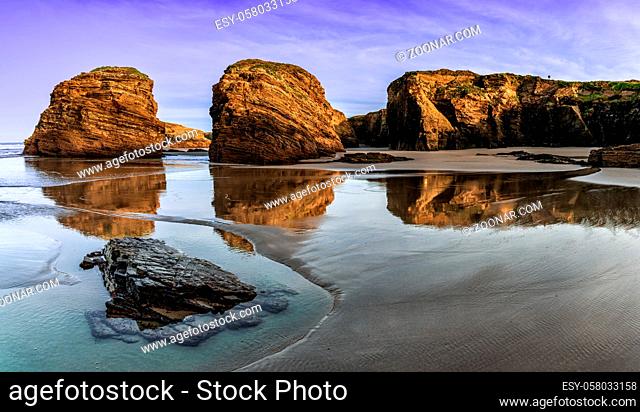 A peaceful sunset on a sandy beach with tidal pools and jagged broken cliffs behind