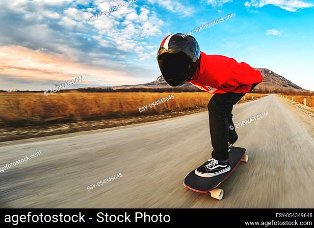 A young man in a fullface helmet and a leather suit in a special rack rides a longboard on afsaltu sunset in the background mountains and beautiful sky at high...