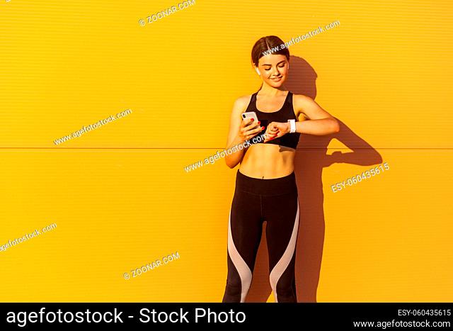 Young attractive girl wearing black sporwear practicing sport exercise in morning on street, standing near orange wall background