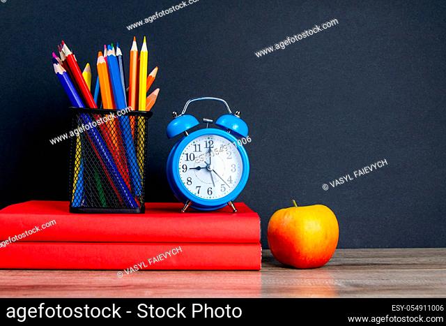 The blue alarm clock along with multi-colored pencils stand on a stack of red books, next to an apple