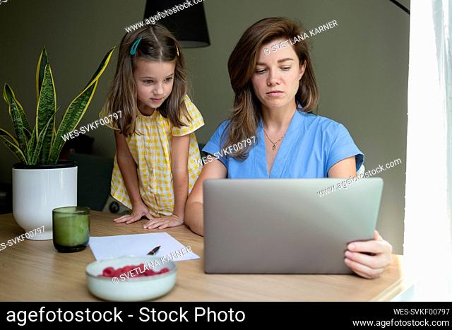 Businesswoman working on laptop with daughter standing by desk at home