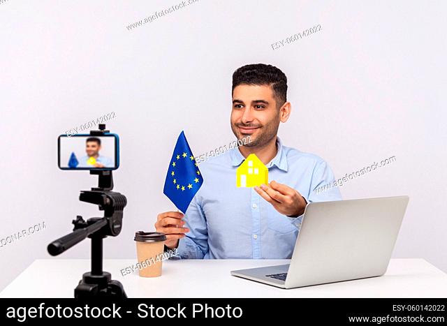 Cheerful smiling man, real estate agent showing paper house and European Union flag to camera, recording video, streaming online blog about purchase or rental...