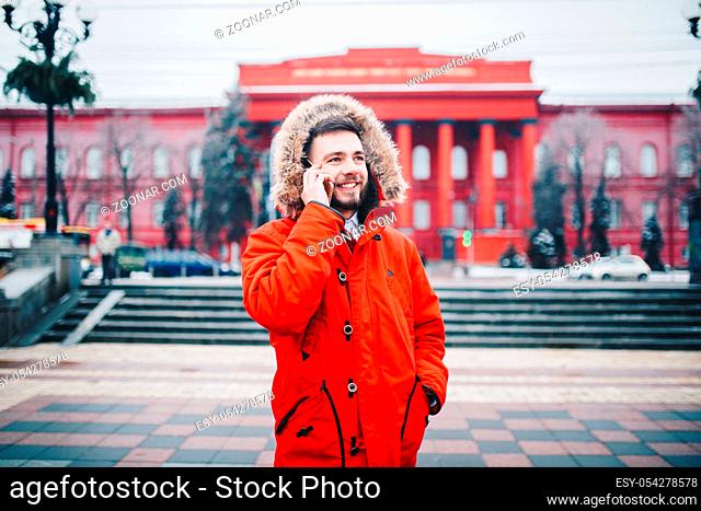 young guy with beard and red jacket in hood a student uses mobile phone, holds in his hand near the head, talking on the phone with smile on the background of...