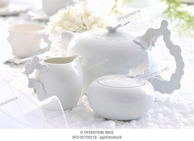 A white tea pot with a sugar bowl and a milk jug on a white table cloth