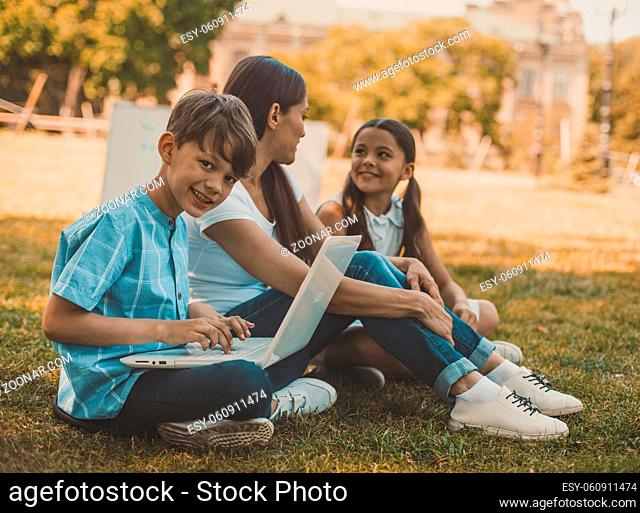 Lesson In Nature, Boy Smiles Looking At Camera And Using Laptop, Teacher Talks With Girl Sitting On Grass Nearby, At Shadow In Sunny Day, Toned Image