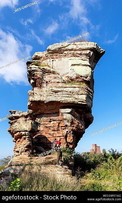 Germany, Rhineland-Palatinate, Senior hiker standing in front of sandstone rock formation in Palatinate Forest with Trifels Castle in distant background