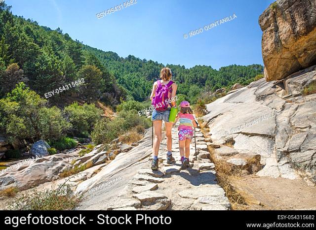 from behind mountaineer family: mother and daughter, five years old girl, with caps and trekking sticks, hiking on stones trail in Camorza Gorge (Madrid, Spain