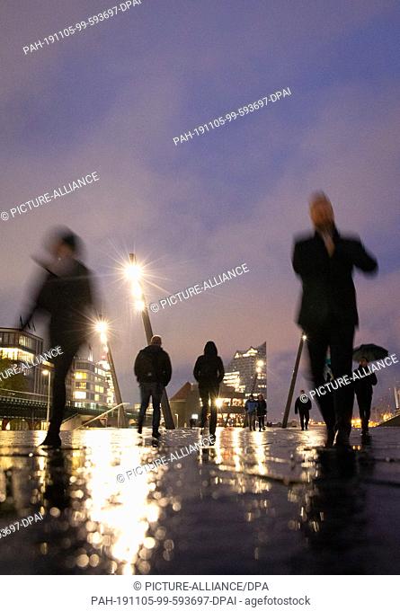 05 November 2019, Hamburg: Passers-by are on their way along the promenade at the landing stages in light rain and dusk. In the background you can see the...