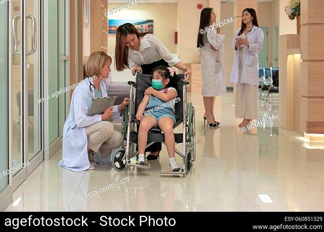 Little girl patient in wheelchair with medical facial mask and mother standing behind in hospital corridor with female doctor kneeling and check up