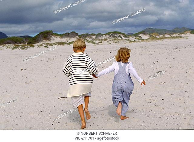 beach-scene, couple viewed from the back, blond girl wearing trousers with braces and blond boy with striped pullover run hand in hand bare feeted through the...