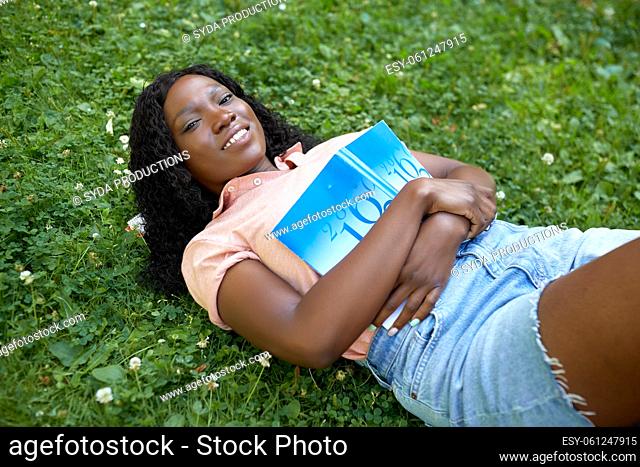 african student girl with textbook lying on grass