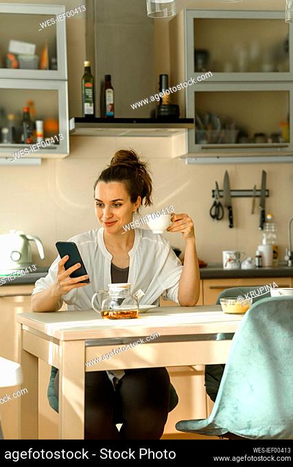 Smiling woman using smart phone and having tea at home