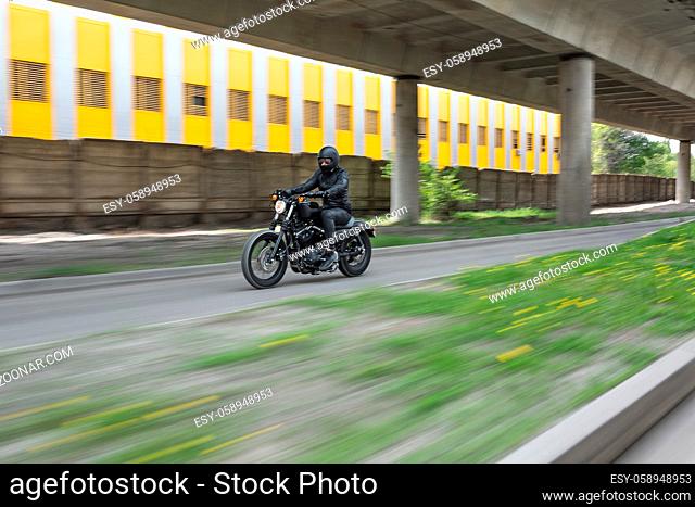 Guy in black helmet hurtling on the black motorbike on the road under the overpass. He wears a black leather jacket, black jeans, black shoes
