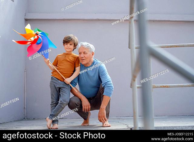 Thoughtful senior man by grandson holding multi colored pinwheel toy in corridor