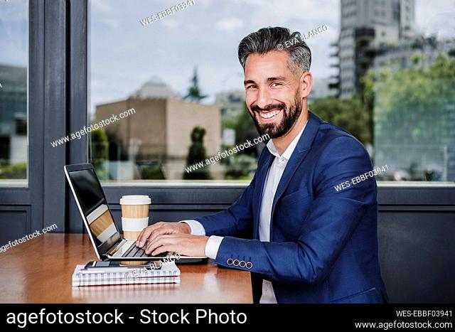 Smiling businessman with laptop sitting by window at cafeteria