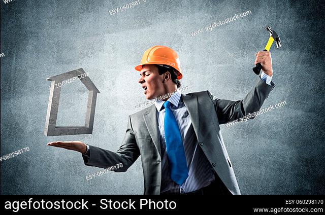Businessman going to crash with hammer house symbol. Young contractor in business suit and safety helmet standing on grey wall background