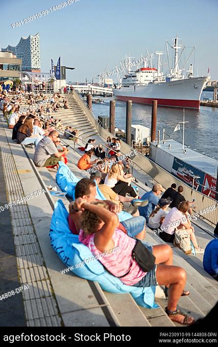 10 September 2023, Hamburg: Numerous people enjoy the evening sun on the Jan Fedder Promenade at the harbor. In the background, the Elbphilharmonie