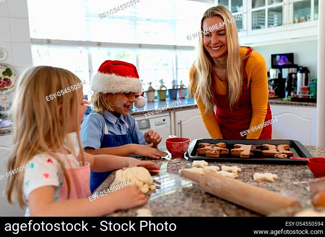 Front view of a happy young Caucasian mother with her young daughter and son in their kitchen at Christmas time making cookies, the children rolling dough
