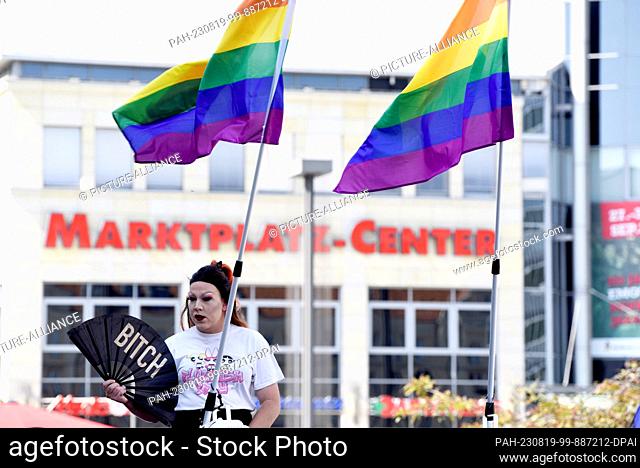19 August 2023, Mecklenburg-Western Pomerania, Neubrandenburg: A participant with a fan stands under rainbow flags on Christopher Street Day