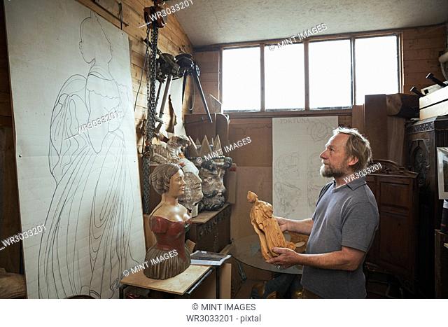 A craftsman wood carver holding a small wooden figure, looking at a large design on the wall of his workshop. Carved wooden ship's figureheads