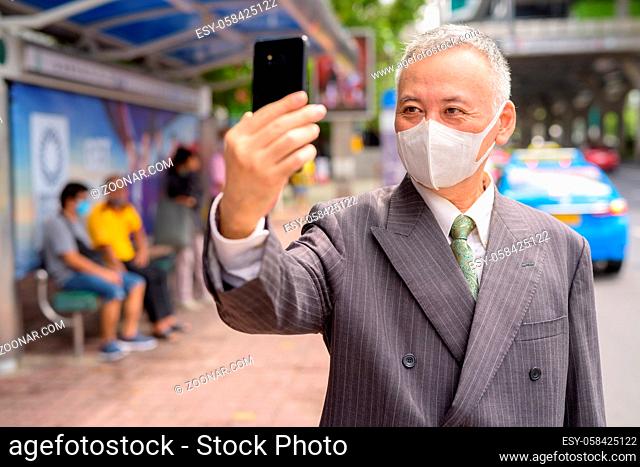 Portrait of mature Japanese businessman with mask for protection from corona virus outbreak at the bus stop in the city
