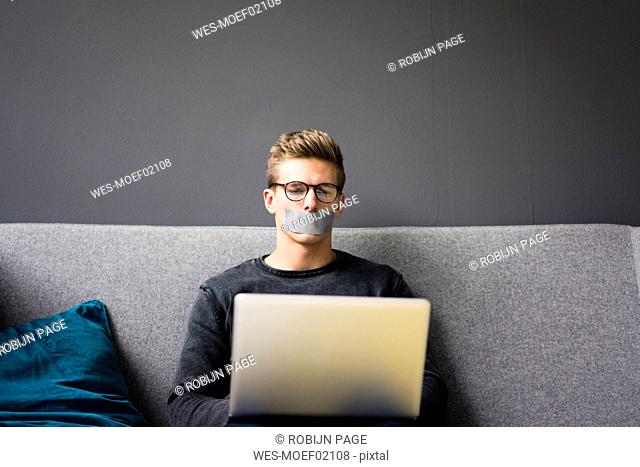 Young man with taped mouth sitting on couch using laptop