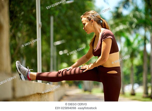 Young blond woman warming up before starting her daily sport routine. The girl does legs stretching in park, near trees. Full length shot