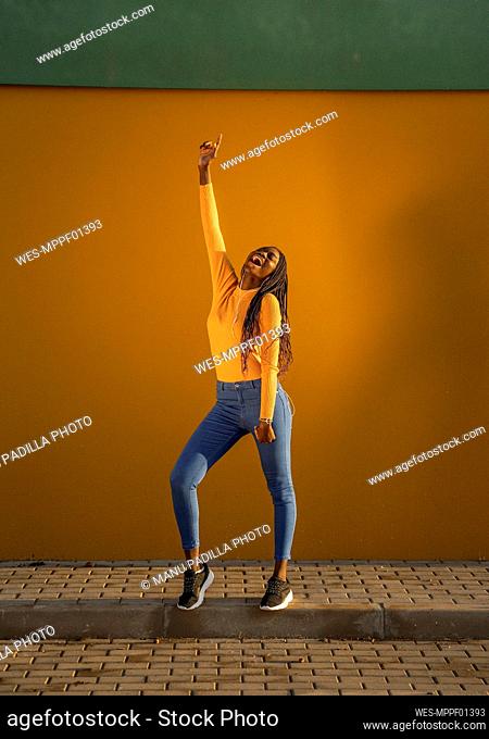 Teenage girl dancing with hand raised against yellow wall on street