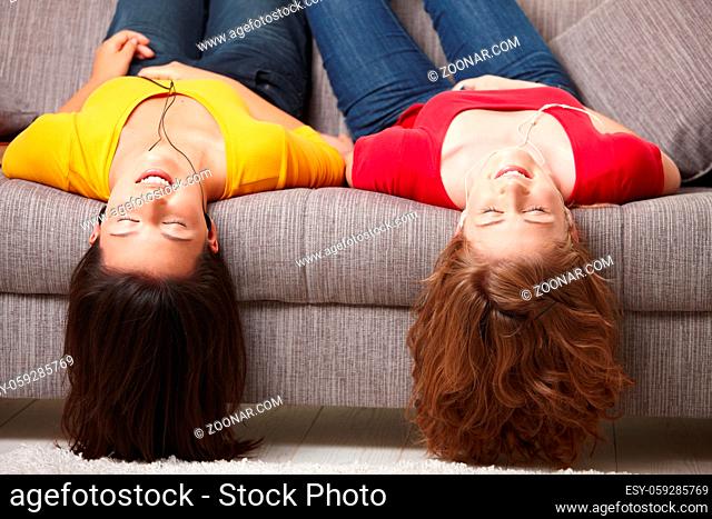 Teen girls lying on couch upside down, listening to music in earbuds, eyes closed