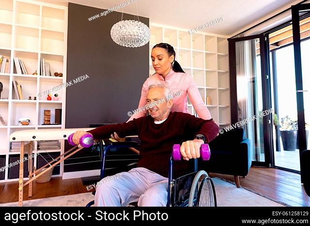 Female biracial physiotherapist assisting caucasian senior man in lifting dumbbells at home