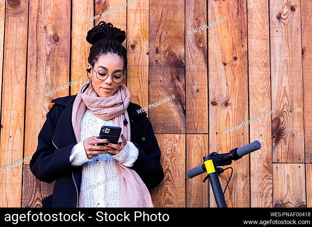 Young female millennial using smart phone while leaning against wooden wall by electric push scooter