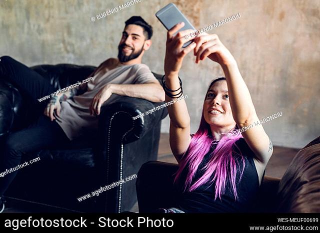 Happy young woman taking selfie with man on sofa in a loft
