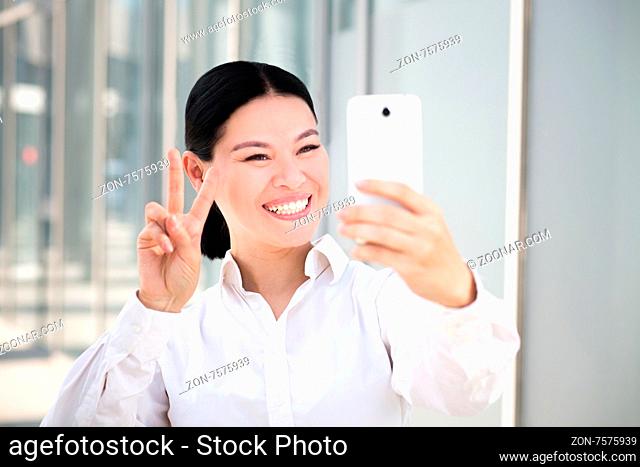 Young businesswoman making selfies on smart phone. Smiling lady in white shirt showing yo sign to the camera on her cell phone