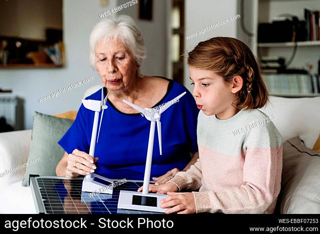 Girl with grandmother blowing model of wind turbine on solar panel at home