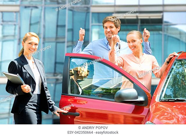 Female Dealer Standing Besides Happy Couple Holding Key Of Newly Purchased Red Car