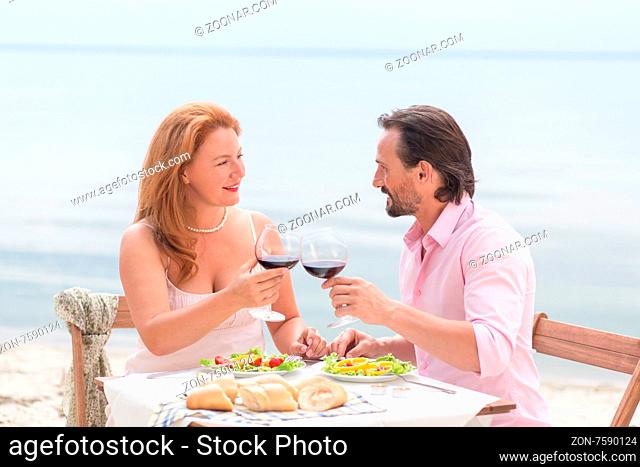 Happy middle-aged couple relaxing at the beach. Man and woman having lunch and drinking delicious red wine near the sea