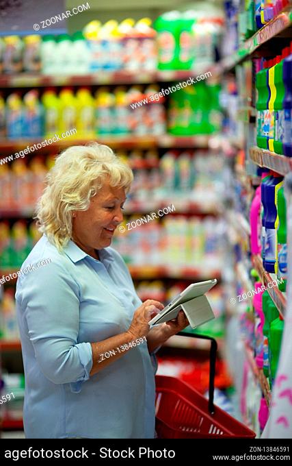 A middle aged fair haired woman is standing in a household section of a supermarket, close to the shelves. She is looking at a tablet screen holding it in hands...