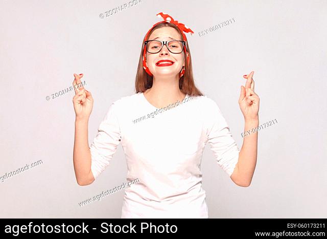 hopeful portrait of beautiful emotional young woman in white t-shirt with glasses, red lips and head band looking at camera with crossed finger