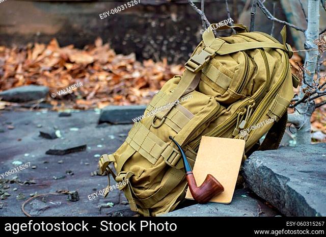 Close up still life style shot of a green army tactical backpack, a paper notebook and a vintage pipe disposed on rock steps in the woodlands