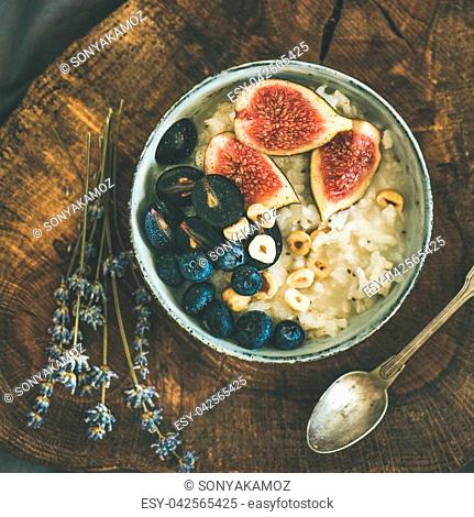 Healthy winter breakfast. Rice coconut porridge with figs, berries and hazelnuts in bowl over rustic wooden board background, top view, square crop