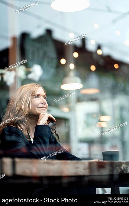 Thoughtful female professional sitting with hand on chin in cafe seen through glass window