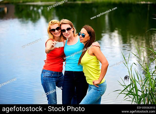 three beautiful young women spending time ourdoors in countryside near lake. friends making phone selfie and having fun. Outside shot. Copy space