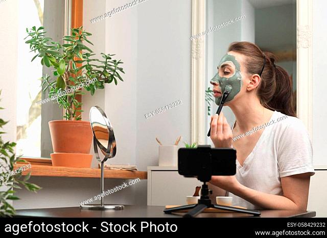 Beautiful young women making online training course about skincare by applying green clay mask by a mirror and in front of smart phone